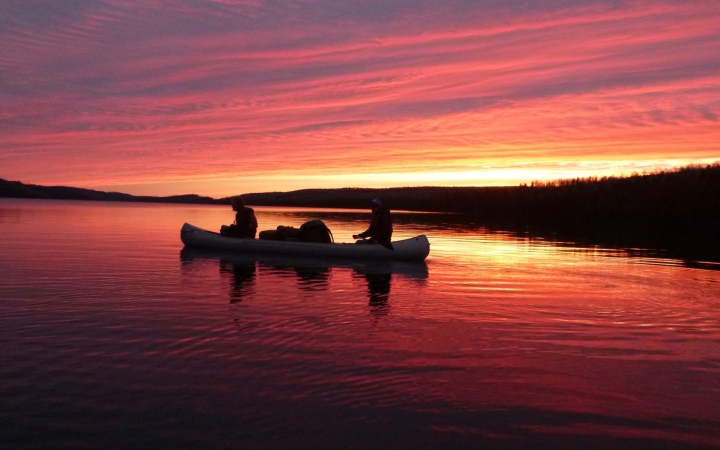 The sky appears in shades of red, orange and purple, reflected in the lake below. On the water, the silhouette of a canoe containing two people can be seen.    